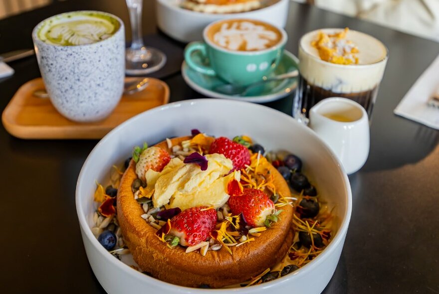 A bowl with a fruit topped souffle pancake, on a cafe table with specialty drinks.