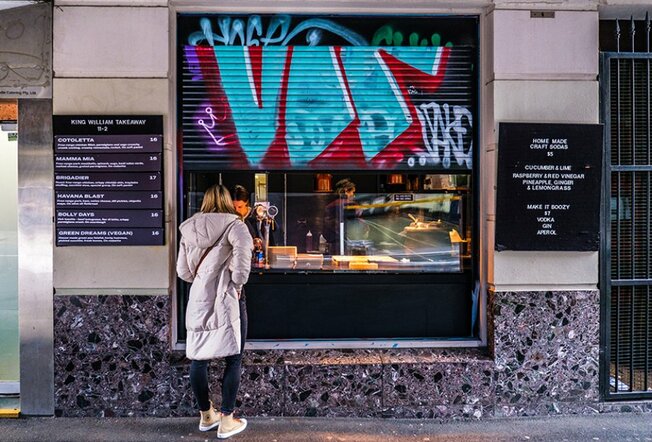 A woman ordering from a hole-in-the-wall cafe behind a graffiti covered roller door.