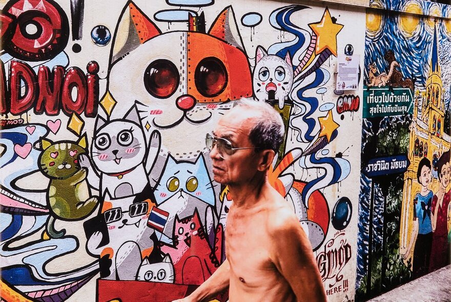 Older man with a naked torso, walking down a street in front of a wall covered in colourful street art and paste-ups.