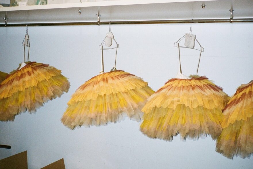 Four feathery like yellow ballet tutus hanging off special hangers against a white wall.