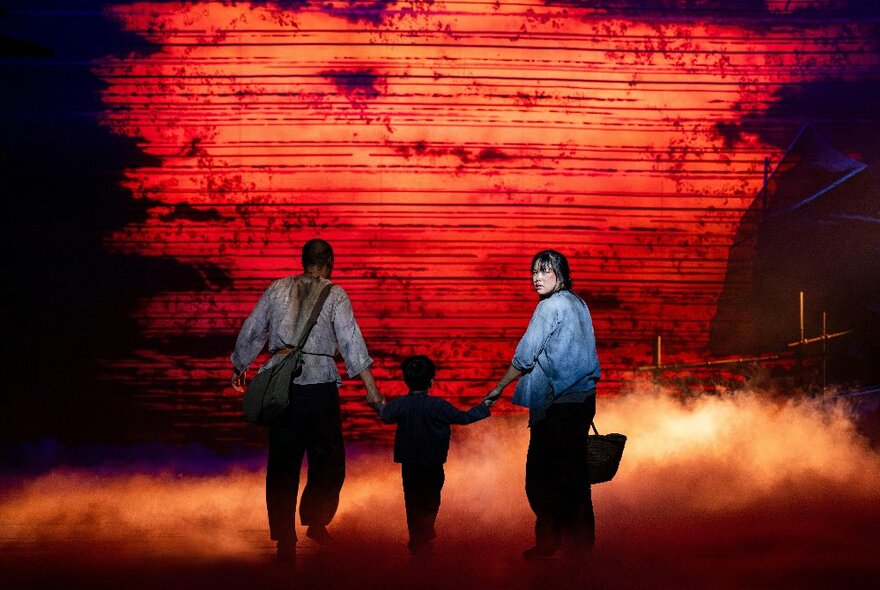 A red coloured theatre stage, two adults and a child walking towards the rear of the stage, the woman looking back over her shoulder at the audience.