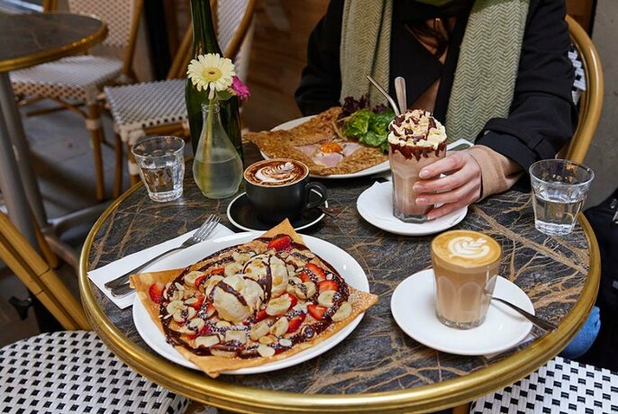 Person seated at a small cafe table set with a chocolate crepe with cream, an iced coffee and a latte.