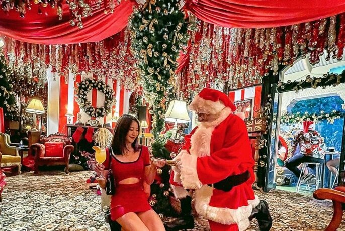 A woman in a red dress and Santa standing in a room decorated with Christmas ornaments, a Christmas tree, baubles and wreaths.