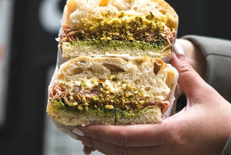 Wrapped focaccia sandwich with creamy white filling and chopped pistachios, held by a person with grey sleeve.