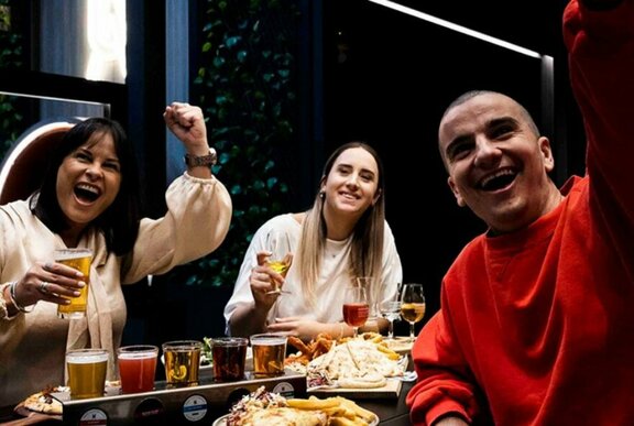 Three people cheering at a table laden with plates of food and pints of beer, in a pub setting. 