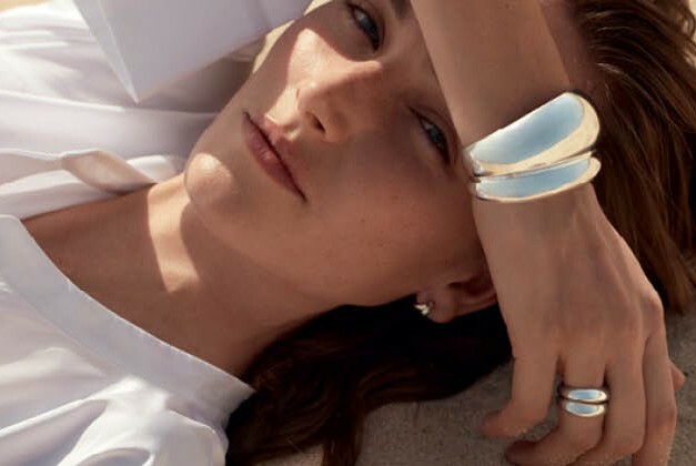 Reclining woman in white with arm over forehead wearing chunky sliver bracelet and silver rings.