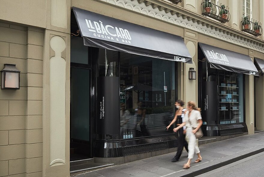 Exterior of Il Bacaro restaurant with two blurred figures walking past on the footpath out the front. 
