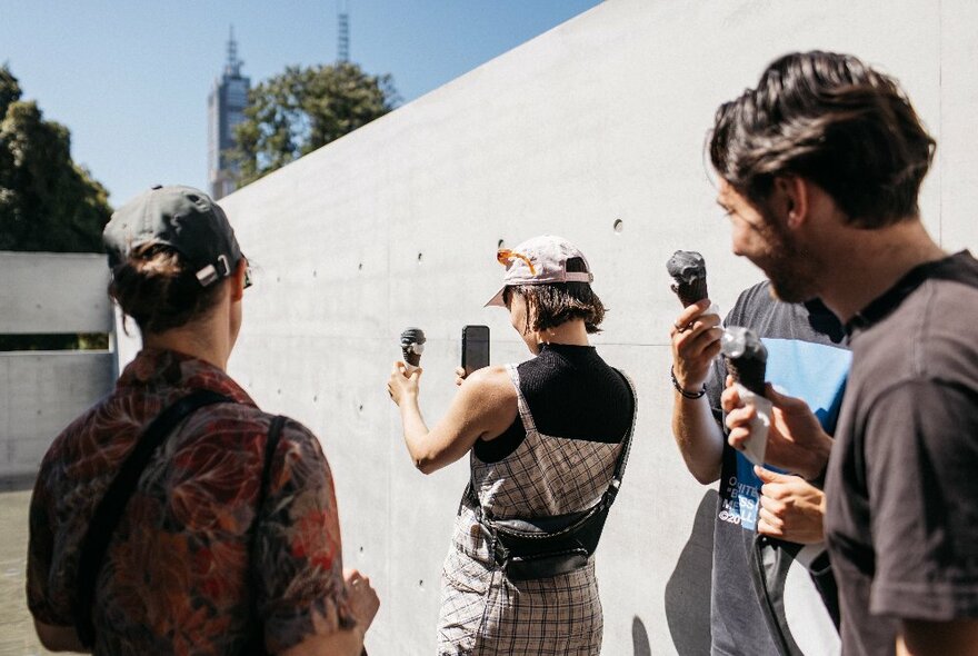 Some people holding grey coloured gelato ice-creams, one taking a photo of theirs held up against a grey concrete wall in an open air structure.