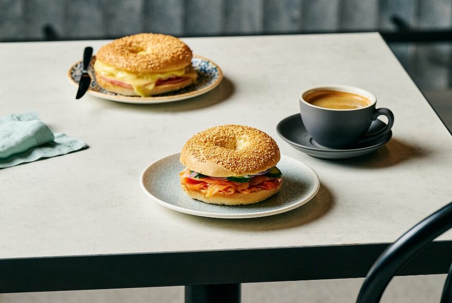 A cafe table set with two filled bagels and a flat white coffee.