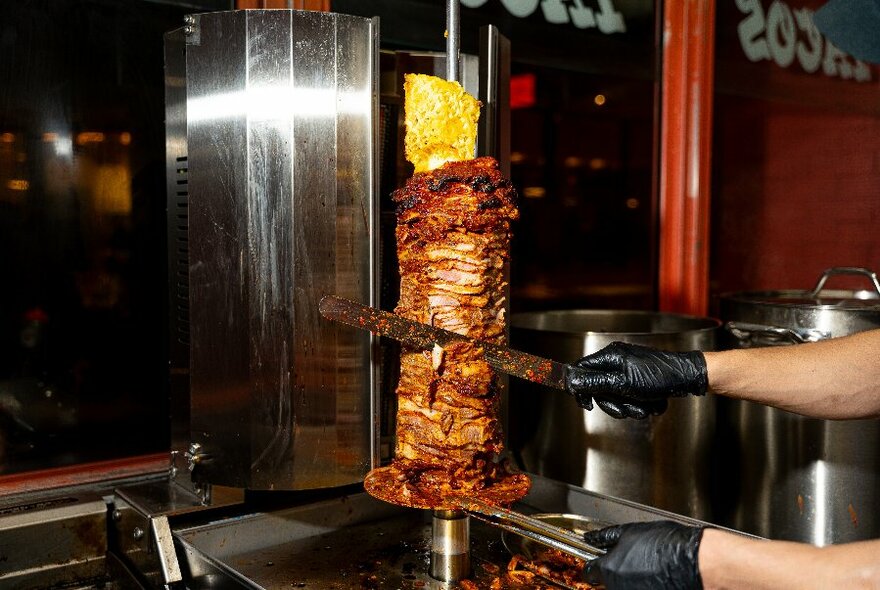 Hands wearing black kitchen gloves slicing meat from a vertical rotisserie grill in a small kitchen setting.