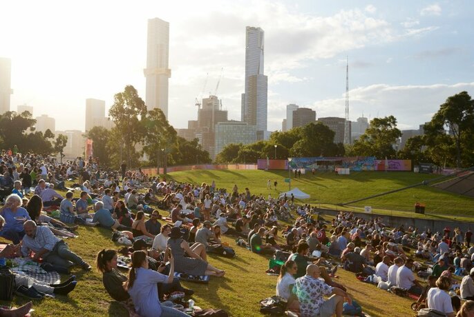 Groups of people relaxing and sitting on rugs on sloping lawns at the Sidney Myer Music Bowl Reserve, with the sun setting in the sky and tall city skyscrapers visible.