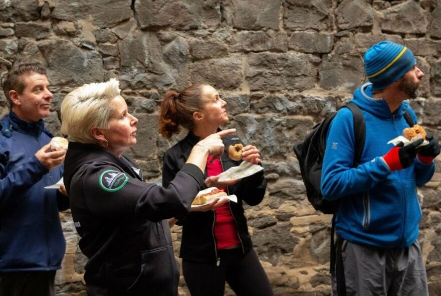 A group of people eating donuts and looking up at something the group leader is pointing to.