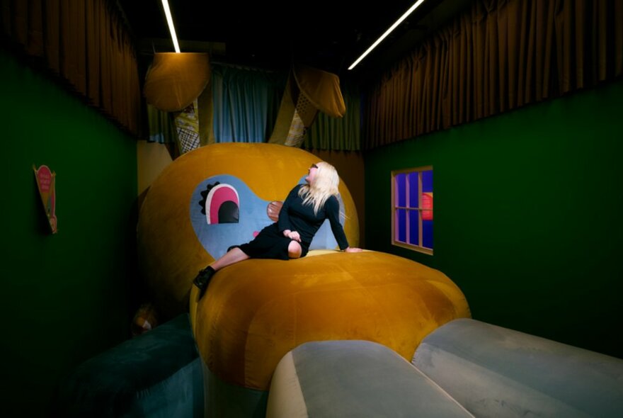 A woman in black, seated on a large velour bunny rabbit in a small green room.