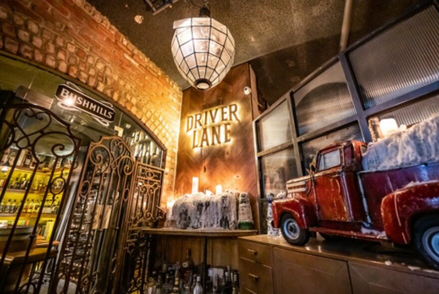 A neon sign inside an old vintage building next to a red toy truck with old candle wax, an old gate and alcohol bottles.