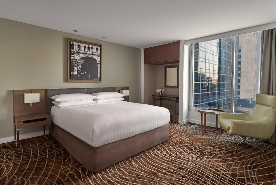 A room at the Melbourne Marriott Hotel with a king- sized bed covered in white linen, a green armchair near large windows and an artwork hanging on the wall above the bed.
