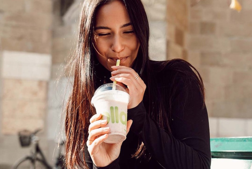 Woman with long dark hair sipping from plastic cup, stone building behind.