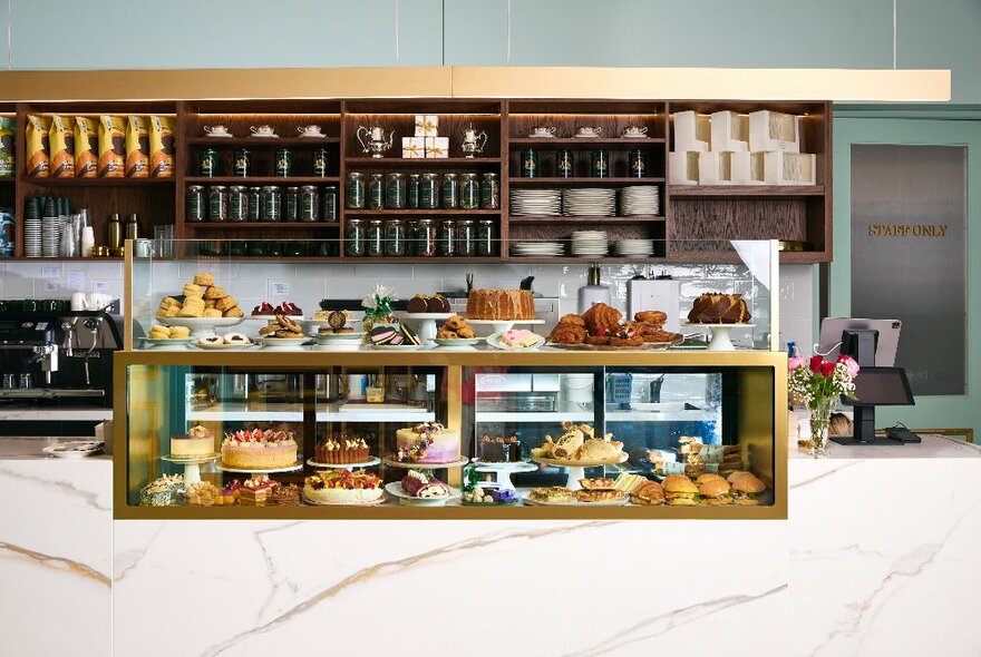 Bakery counter with cakes and desserts on plates in a gold-edged counter-top display case, with shelves of cafe goods in the background.