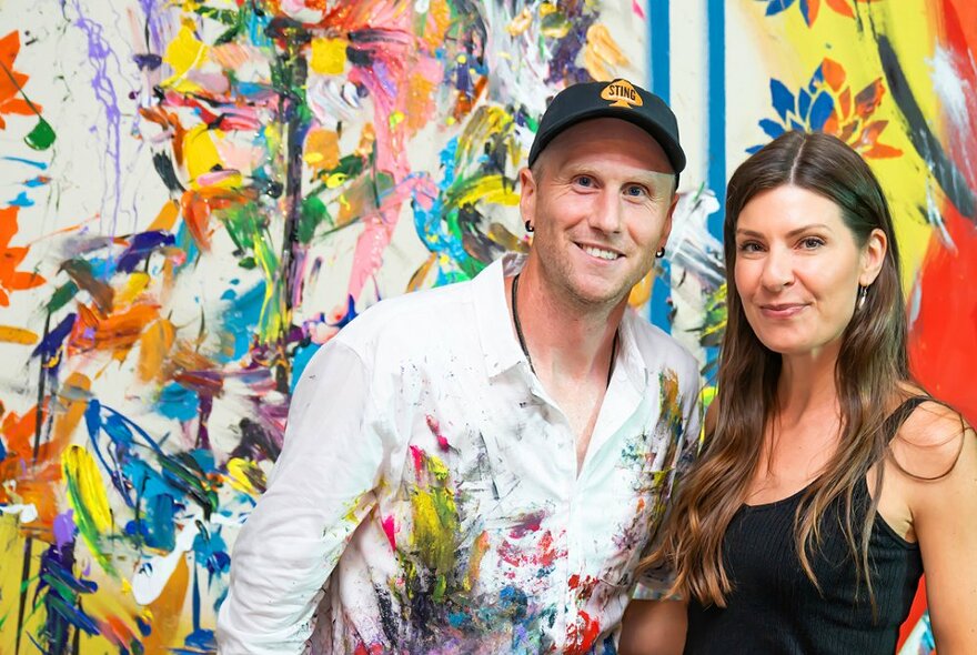 A man and woman pose in front of a graffiti-like painting, the paint is also on the man's shirt. 