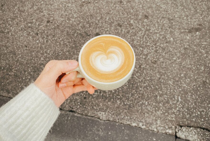 A hand holding a white cup of caffe latte against concrete.