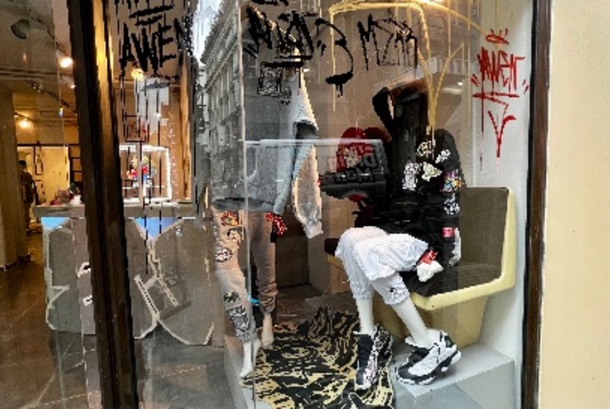 A store window showing a mannequin seated in a black hoodie with graffiti on the window.