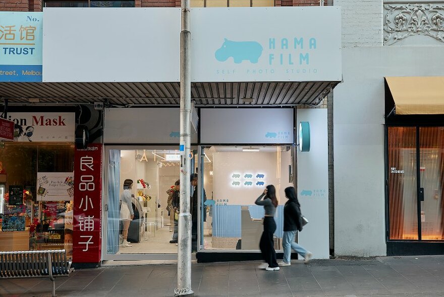 Two girls walking into a white and blue store called Hama Film.