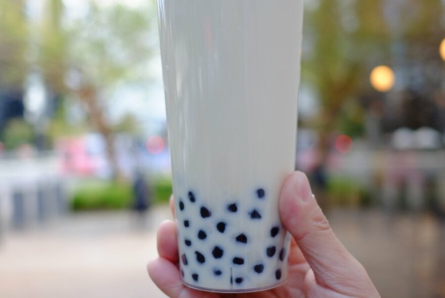 Cream-coloured drink with seeds at bottom, in plastic cup, held up by hand, blurred cityscape with trees behind.