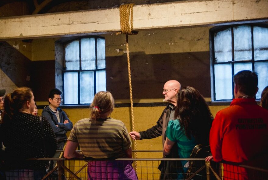 A tour leader holds a hangman's rope on a beam, with visitors listening and looking on.