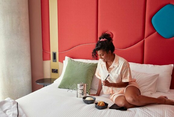 Woman seated on a hotel bed in shortie pyjamas eating tiffin dishes of rice and curry.