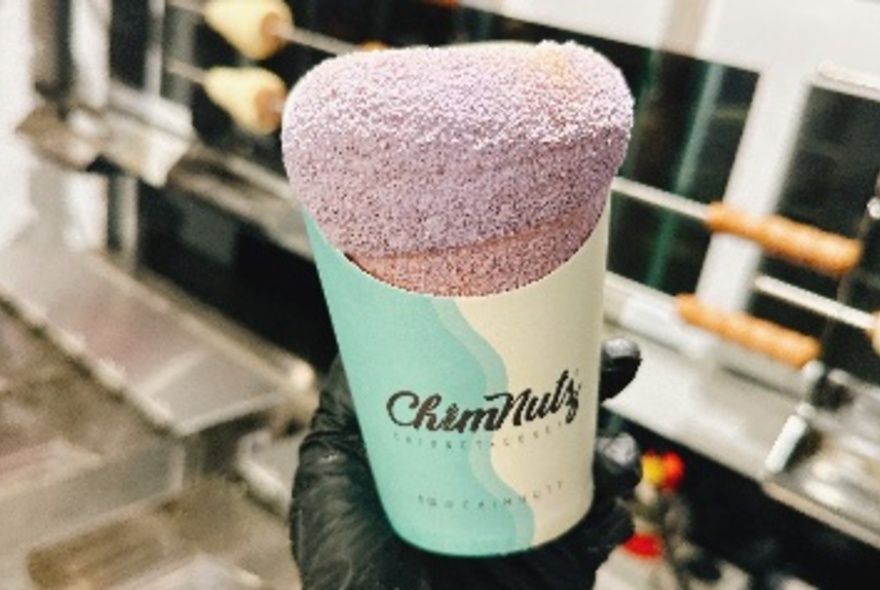 A chimney cake in a paper holder with lavender coloured sugar coating.