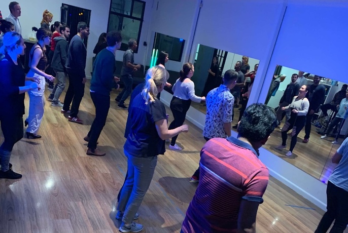 An angled image of people dancing in pairs, facing towards mirrored wall, in a blue-lit room.