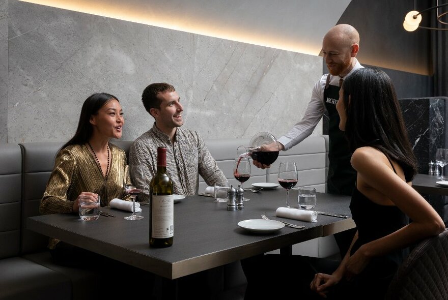 Waiter pouring red wine from carafes with three people at a table in a contemporary setting.