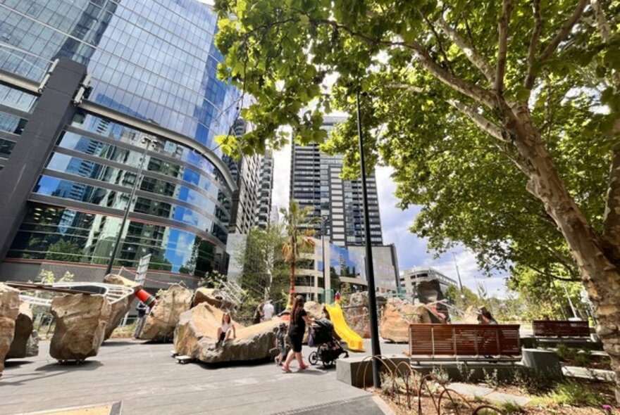 Park made from natural elements, boulders and a yellow slide. City skyscrapers in the background with a big tree.