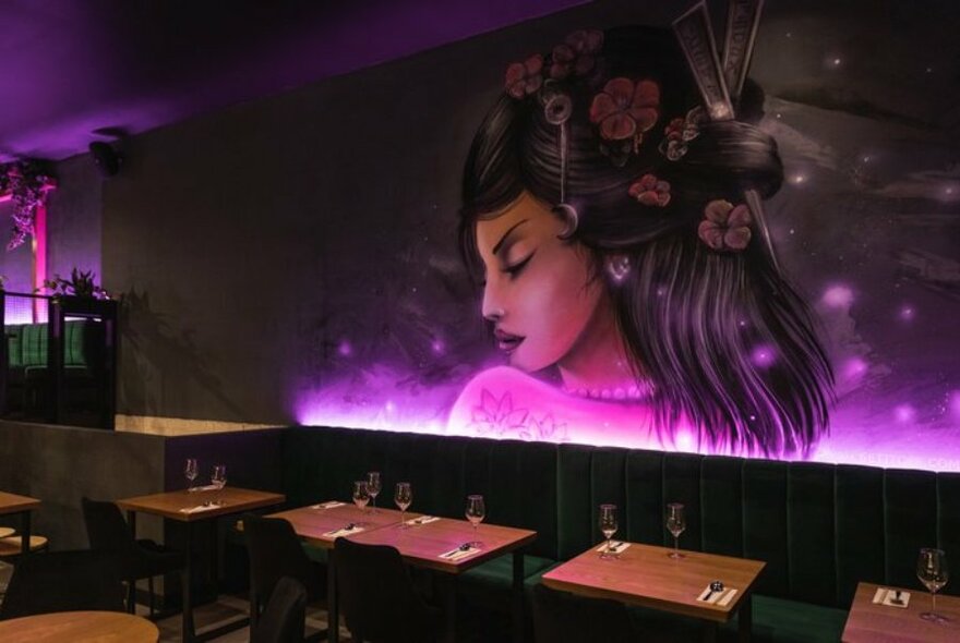 A woman's face in profile with flowers in her hair painted on a wall in a restaurant with a pink glow. 