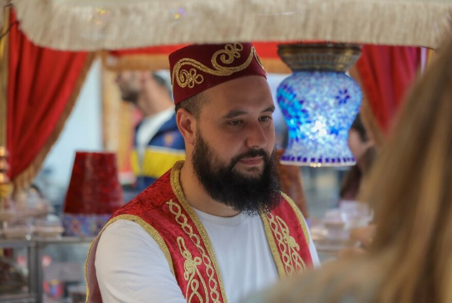 A man wearing a traditional Turkish red embroidered fez and a matching waistcoat, at a stall selling Turkish handicrafts.