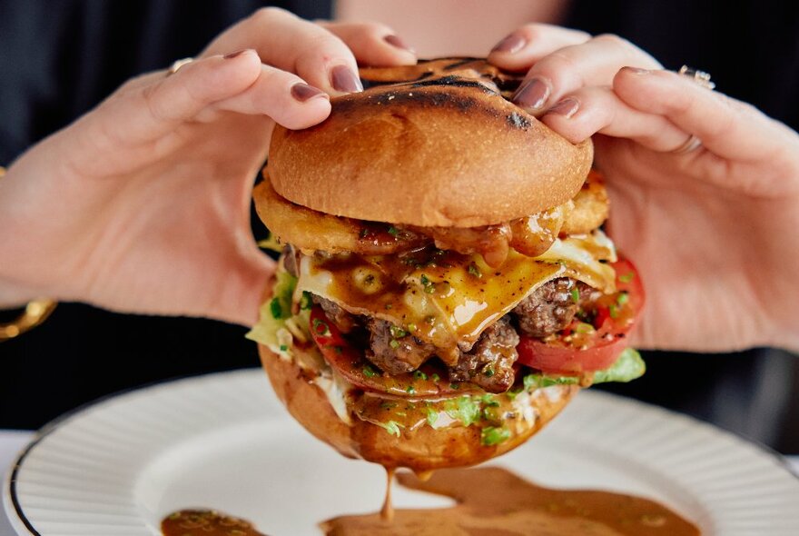 Two hands holding up a very sloppy burger with sauce dripping onto a plate. 