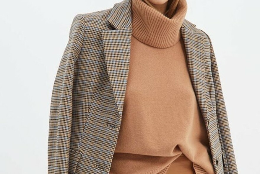 Model wearing a brown turtleneck jumper and draped plaid jacket.