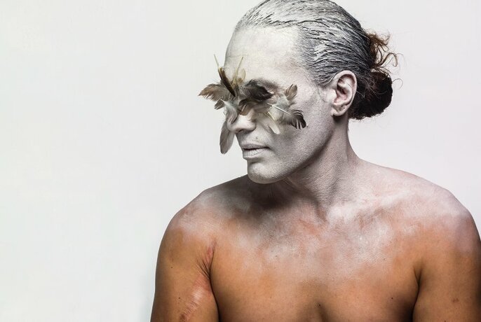 Half bust torso of a naked man, his face and pulled-back hair painted white, with silvery grey bird feathers covering his eyes, posing against a white background.