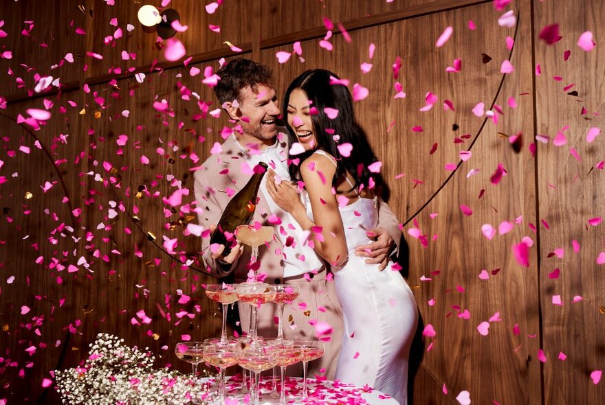 A bride and groom hugging and celebrating in a room, in front of tower of champagne glasses the groom fills from a bottle of pink sparking wine, with pink confetti all around them.