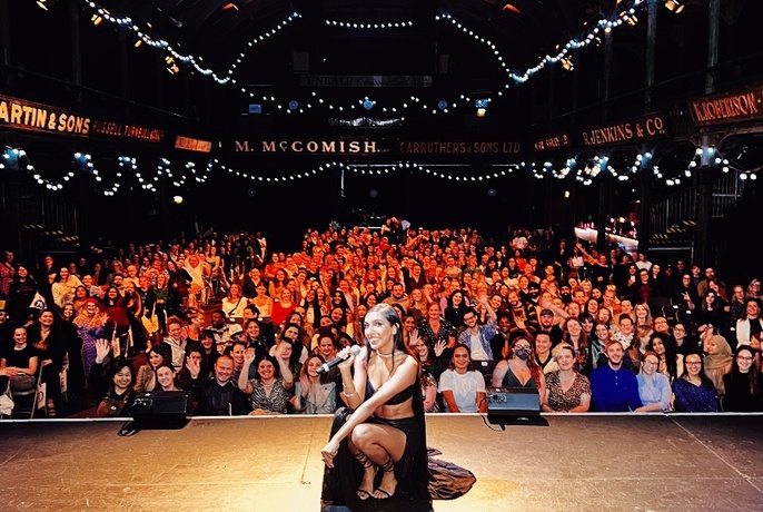 Poet Rupi Kaur smiling and squatting down on a theatre stage with her back to a large seated audience behind her.