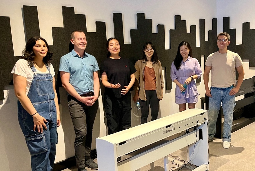 A group of people inside a music studio, smiling and standing in front of a digital piano keyboard.