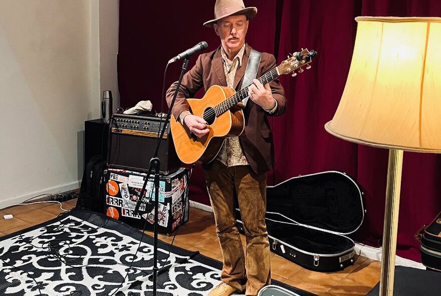 Dave Graney doing an in-store performance at One Star Gallery, playing acoustic guitar standing against red curtains next to a yellow standard lamp.