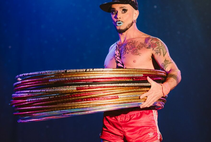 A tattooed circus performer wearing red footy shorts, a cap and blue lipstick, holding a large number of hula hoops around his waist.