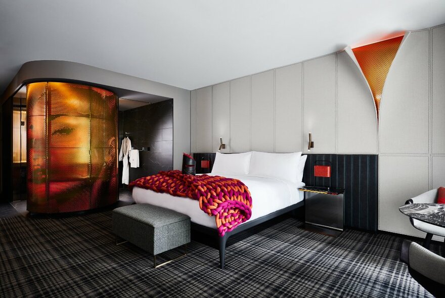 Contemporary hotel room with feature decor including gold-reveal wallpaper and bathroom pod, checked carpet and oversized pink knit blanket.