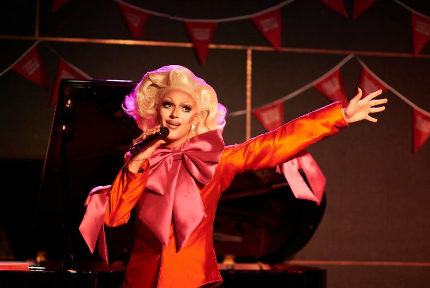 Drag artist on stage, waving arms and wearing a blonde 1950s wig and orange dress with an oversized bow.