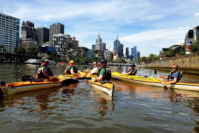 A group of kayakers assembled in a circle on the water with the Melbourne skyline in the background. 