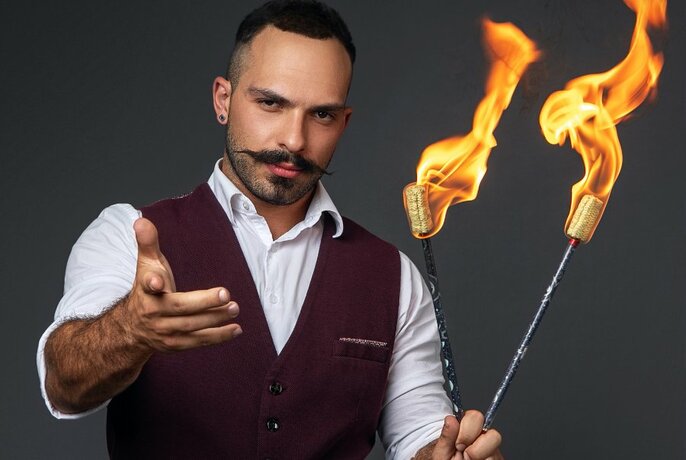 A man with a waxed moustache holding two flaming sticks in one hand while holding his other arm out straight towards the camera.
