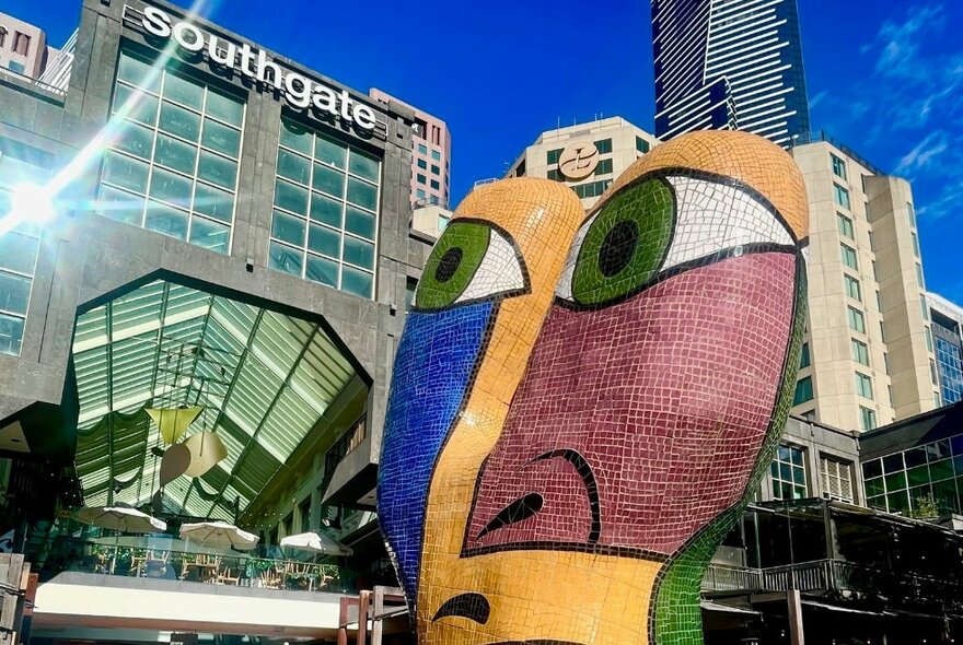 A large outdoor mosaic sculpture of an abstract face, in front of the entrance to Southgate, with several tall buildings in the background.