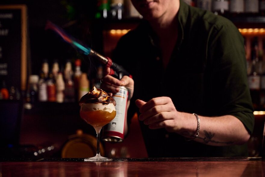 A bartender about to apply a blowtorch to the top of an elaborate cocktail. 