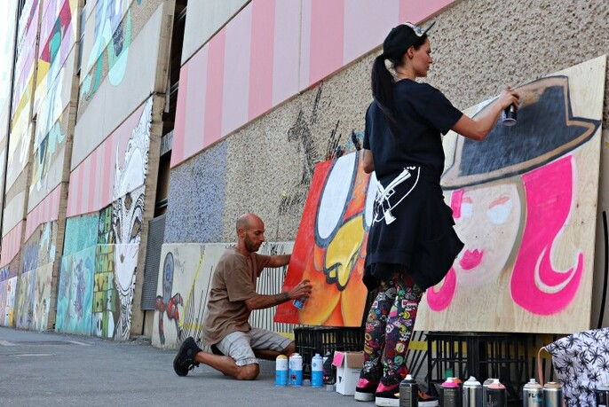Street artists with spray cans creating art on oversized wooden panels in a laneway.