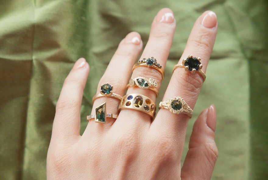 Hand against green background, wearing a number of gold rings with green stones.